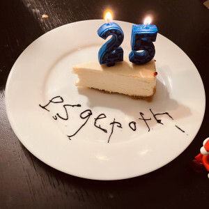 A slice of cheesecake with a 2-shaped and 5-shaped candle and the word esgeroth written below it in icing