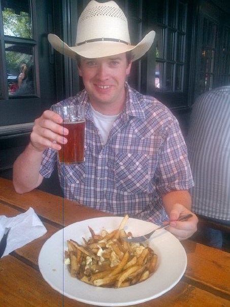 Pete in a cowboy hat holding a beer and eating poutine
