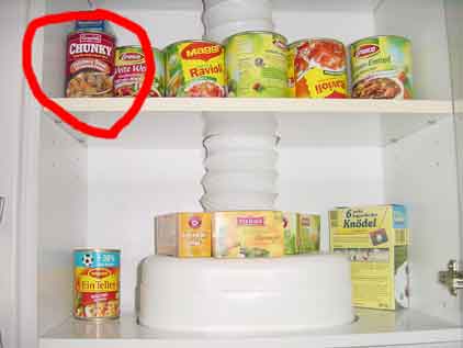 A kitchen cupboard with various cans of food with German labels in it. A single can with an American label "Chunky" is highlighted with a red circle