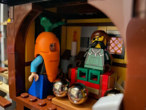 Two Lego minifigures within a room in a Lego castle. One is Princess Anna from Frozen, but wearing a carrot outfit. The other is seated on a chair and has a large mustache