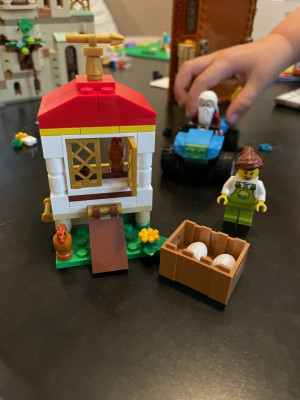 Lego chicken coop with two chickens and a box with eggs. A farmer minifigure stands near the box. In the background a child's hand is pushing a Lego four-wheeler with Dumbledore in the driver's seat