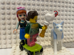 Lego skateboard with two minifigures on it. In the front is a minifigure in pink pants and a green shirt, holding a chicken. In the back is Anna from Frozen. Next to them is a large Lego dog.